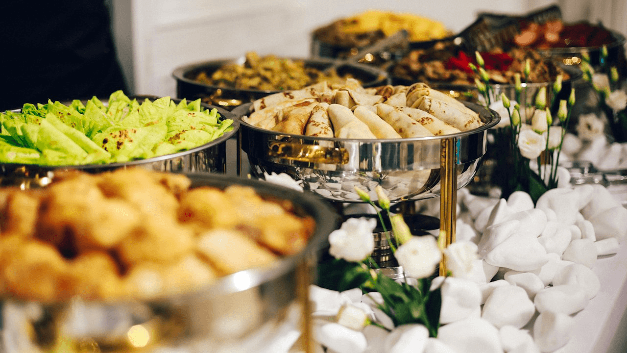 Mesa de buffet real com rechauds de prata, salgados dourados e wraps, luz natural quente, sem texto, logos ou paleta rosa e azul forçada.